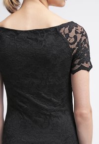 Černé krajkové šaty s designem bez ramínek, s krátkými rukávy a texturovaným květinovým vzorem na látce.