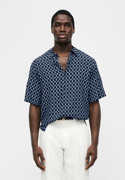 Moški stoji, oblečen v temno modro kratko srajco s svetlomodrim okroglim vzorcem in belimi hlačami na enobarvnem ozadju.