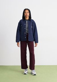 Giacca in denim navy con tasche frontali, abbinata a una maglietta a righe e pantaloni in corduroy bordeaux. Sneakers grigie completano l'outfit.