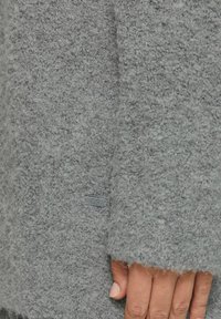 Grauer, flauschiger Pulloverstoff mit weicher Textur. Merkmale sind ein auffälliges graues Etikett, sichtbare Nähte und locker sitzende Ärmel.