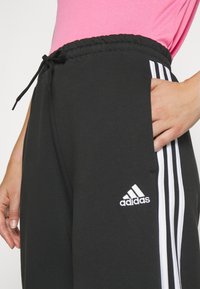 Černé tepláky vyrobené z měkké tkaniny, se třemi bílými pruhy podél boků a vyšitým logem Adidas na stehně.
