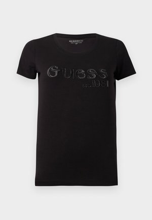 T-shirt noir en coton avec des manches courtes et un col rond. Présente le texte "Guess est. 1981" en lettres en relief ornées de clous argentés.