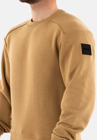 Felpa marrone con collo a giro, maniche lunghe e polsini a coste. Presenta un logo in tessuto nero sulla manica sinistra. Texture liscia.