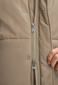 Beżowa kurtka puchowa z centralnym zamkiem, srebrnymi elementami i pikowaną fakturą. Zawiera zatrzaski oraz zbliżenie na dłoń.