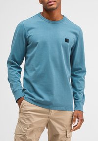 Blaues Langarmshirt aus weichem Stoff, mit rundem Halsausschnitt und einem kleinen schwarzen Logopatch auf der linken Brust. Kombiniert mit beigen Cargo-Hosen.