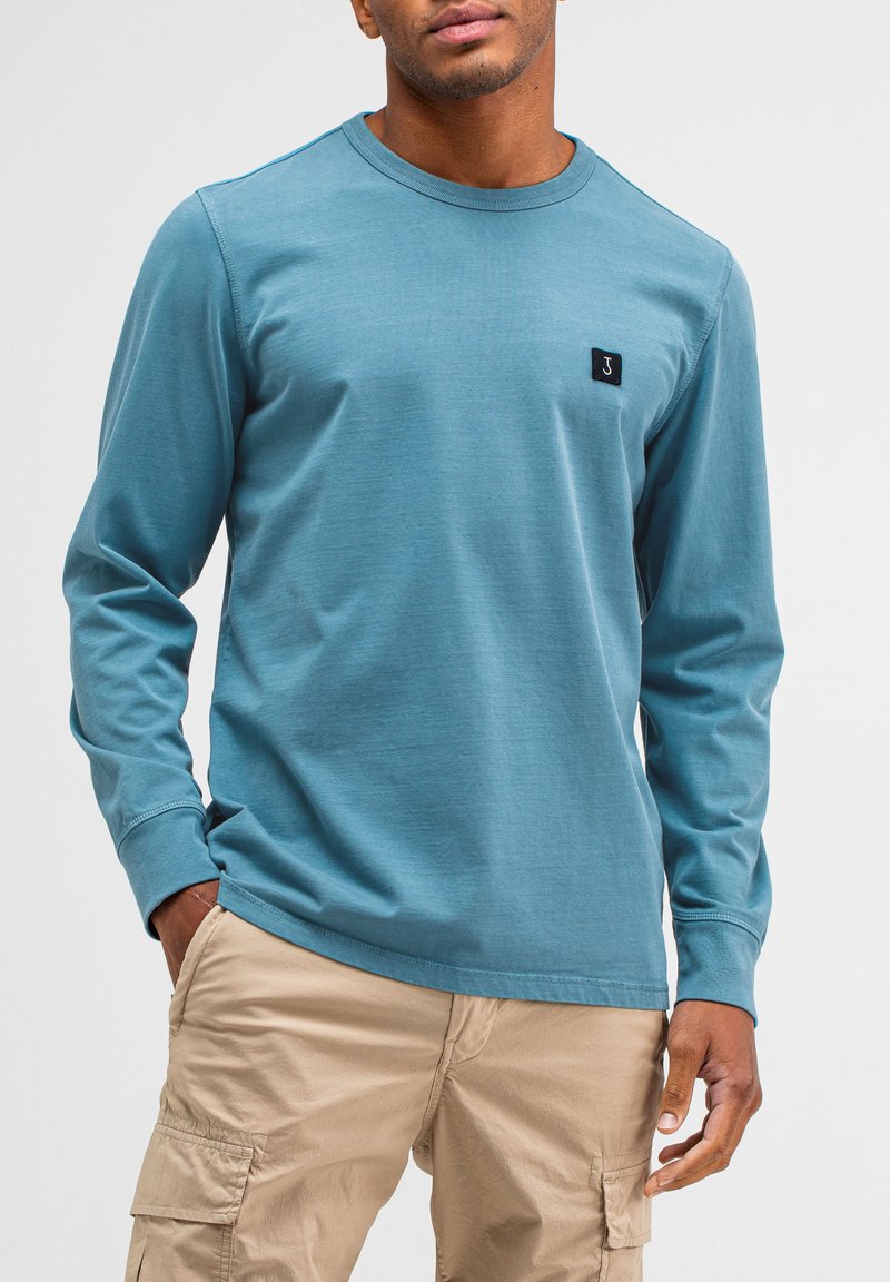 Blaues Langarmshirt aus weichem Stoff, mit rundem Halsausschnitt und einem kleinen schwarzen Logopatch auf der linken Brust. Kombiniert mit beigen Cargo-Hosen.