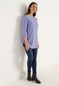 Kvinna som står mot en vit vägg klädd i en ljuslila långärmad topp, mörkblå tajta jeans och svarta ankelboots.