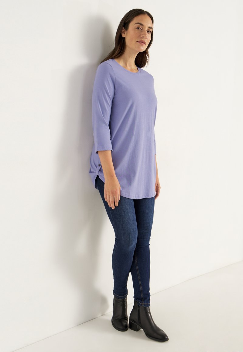 Kvinna som står mot en vit vägg klädd i en ljuslila långärmad topp, mörkblå tajta jeans och svarta ankelboots.