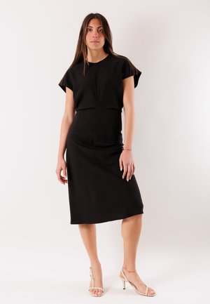 Donna in piedi che indossa un abito midi nero a maniche corte e tacchi aperti beige su uno sfondo bianco semplice.