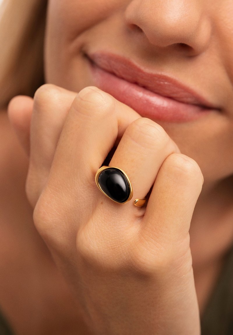 Anello d'oro con una pietra ovale nera, superficie liscia e una fascia sottile, indossato su una mano con pelle chiara, vicino a uno sfondo neutro.