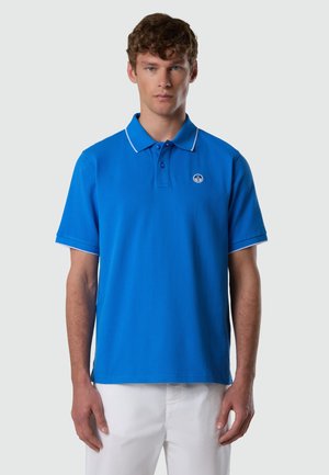Homme portant un polo bleu à manches courtes avec des bordures blanches et un pantalon blanc, debout devant un fond gris clair uni.