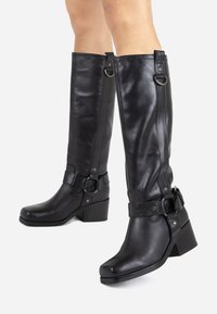 Bottes montantes en cuir noir avec un embout carré, un talon bloc, des sangles décoratives et des accents en métal. Texture lisse et design ajusté.