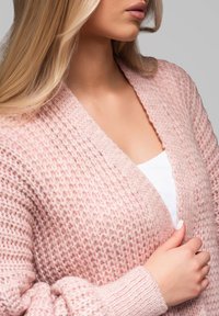 Světle růžový pletený cardigan s texturovaným vzorem a otevřeným předním dílem. Má dlouhé rukávy a žebrované manžety, nošený přes bílý top.