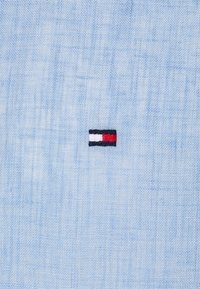Tissu texturé bleu clair avec un petit logo brodé rectangulaire comportant des sections horizontales blanches, rouges et bleu marine.