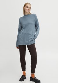 Pull bleu à manches longues avec un col montant, texture douce, associé à un pantalon fuselé marron et des mocassins noirs. Des chaussettes orange ajoutent une touche de couleur.