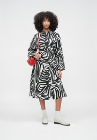 Juoda ir balta dryžuota suknelė su suspaustu juosmeniu, ilgomis rankovėmis ir laisva silueta. Derinama su maža raudona rankine ir baltais batais.