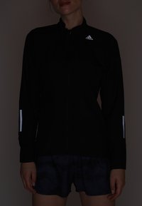 Veste de sport noire avec fermeture éclair complète, bandes réfléchissantes sur les manches, texture lisse, associée à un short bleu foncé à motifs.