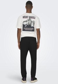 Witte t-shirt met een afbeelding van de Makalu op de rug, zwarte broek en zwarte schoenen met paarse accenten. Relaxte pasvorm, korte mouwen.