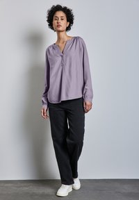 Lila långärmad blus med V-hals, med gathered axlar. Kombinerad med lösa svarta jeans och vita sneakers på en grå bakgrund.