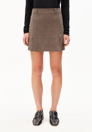 Persoon met een bruine corduroy minirok, zwart langemouwen shirt en zwarte loafers met zilveren studs, staand tegen een witte achtergrond.