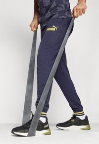 Zapatillas deportivas negras con un acento amarillo, pantalones de chándal azul marino con un logo dorado y una banda de resistencia gris sostenida en la mano.