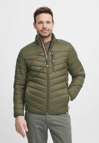Olivegrüner gepolsterter Mantel mit hohem Kragen, diagonaler Steppnaht und kontrastierenden Reißverschlüssen. Seitentaschen sorgen für Funktionalität.