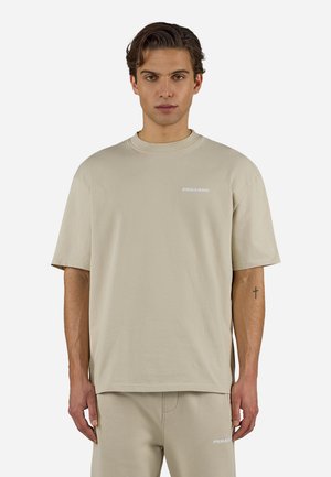 Mann som har på seg beige kortermet T-skjorte og matchende bukser med små hvite "PEGADOR"-logoer, stående mot en ensfarget lys bakgrunn.