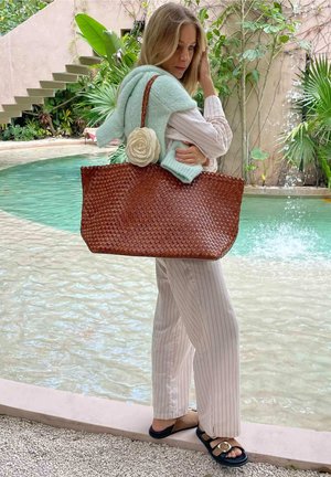 Femme avec un grand sac tressé marron orné d'une rose blanche, portant un pyjama rayé et un pull menthe, debout près d'une piscine avec des plantes et des escaliers.