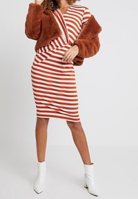 Pruhované bodycon šaty v červené a bílé barvě, doplněné hnědým kožíškem z umělé kožešiny. Modelka má na sobě bílé kotníkové boty.