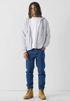 Sudadera gris con cremallera sobre una camiseta blanca, combinada con jeans azules y botas de trabajo color beige. Tejido suave y corte relajado con bolsillos frontales.