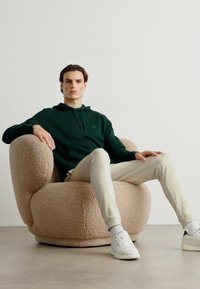 Felpa verde con cappuccio e cordino, pantaloni morbidi crema e sneakers bianche. Seduto su una poltrona rotonda, soffice, di colore beige con una finitura in tessuto texturizzato.