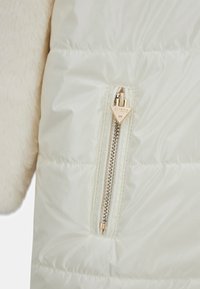 Giubbotto puffa avorio con tessuto morbido simile alla pelliccia sulle maniche, zip dorata con logo triangolare GUESS e texture trapuntata.