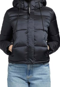 Doudoune noire avec capuche et cordons, portée avec un jean bleu clair, mains dans les poches de la veste, fermeture éclair frontale visible.
