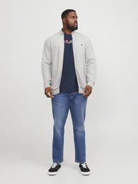 Chaqueta de cierre vertical gris claro sobre una camiseta navy con logo rojo, combinada con jeans azules y zapatillas negras con cordones blancos. Diseño simple y ajustado.
