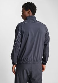 Mörkgrå sportjacka med hög krage, elastiska cuffs och midja. Har kontrasterande ljusgrå sömmar längs axlarna.