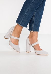 Ljust grå högklackade skor med spetsig tå, tillverkade av mocka, med ett band över vristen och en blockklack.