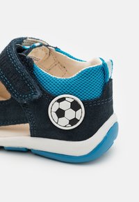 Modrá a černá semišová dětská bota s texturovaným síťovým svrškem, suchým zipem a akcentem v podobě kulatého fotbalového míče na boku.