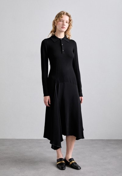Vestito lungo nero a maniche lunghe con colletto polo, orlo asimmetrico e corpetto testurizzato. Abbinato a mocassini neri con dettagli in dorato.