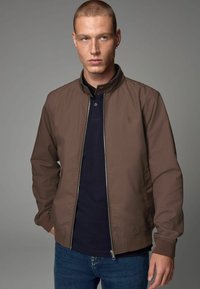 Veste bombardier marron avec fermeture éclair, poignets et col côtelés. Portée sur un polo bleu marine, avec un logo discret sur la poitrine.