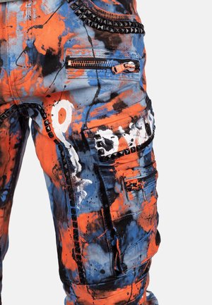 Stof broek met een combinatie van oranje, blauwe en zwarte spetterpatronen. Inclusief ritszakken en zwarte studs langs de naden.