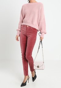 Kobieta ubrana w różowy sweter dzianinowy i mauve welurowe spodnie, trzyma różową torebkę w wzory i nosi czarne, spiczaste szpilki.