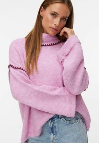 Pull léger rose avec un col haut, présentant une finition texturée et une bordure contrastante rose foncé le long des bords. Silhouette ample.