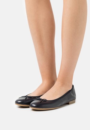 Jambes portant des ballerines en cuir noir avec de petits nœuds et des semelles marron clair, sur un fond blanc uni.