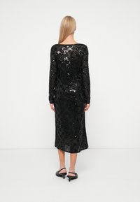 Vestido preto com lantejoulas, mangas longas, comprimento até ao joelho, superfície texturizada, ajuste justo e silhueta reta. Usado com sapatos de salto com tira no tornozelo pretos.