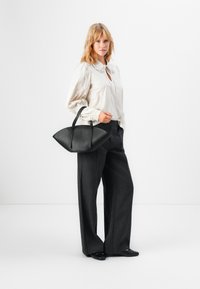 Svart skinnveske med buet, bred form og korte håndtak. Modellen har på seg en kremfarget bluse med volangkrage og grå vide bukser.
