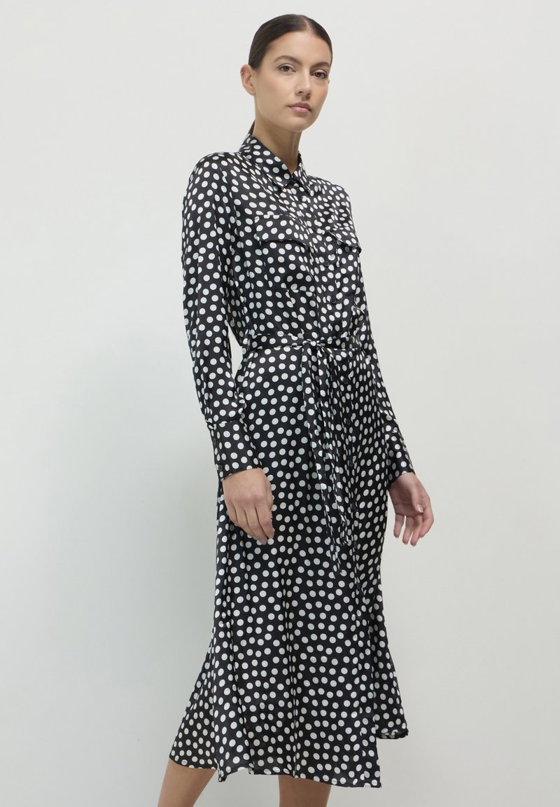Schwarzes Kleid mit weißen Punkten, langen Ärmeln, durchgehender Knopfleiste, zwei Brusttaschen, tailliertem Gürtel und ausgestellt geschnittenem Saum. Glatte Textur.