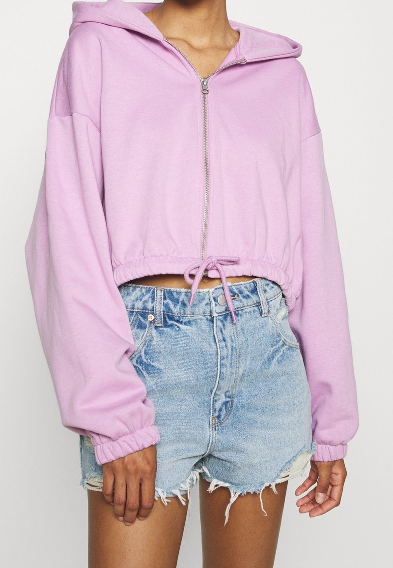 Sweat à capuche court rose clair avec une fermeture éclair frontale et un cordon de serrage, doté de larges manches et d'une texture douce, accompagné de shorts en jean déchirés.