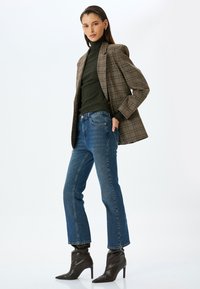 Blazer de cuadro marrón sobre un cuello de tortuga verde oscuro, combinado con jeans rectos azules y botas de tobillo de tacón oscuro. Textura suave en toda la prenda.