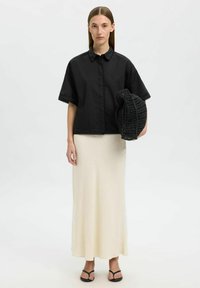 Chemise noire à manches courtes avec dentelle, longue jupe crème clair, sac tissé noir et tongs noires. Fond neutre.
