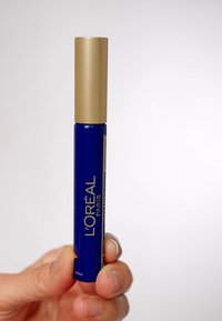 Blå cylindrisk mascaraflaska med ett guldlock, med "L'Oréal Paris" tryckt i guldbokstäver. Hållen i en hand för visning.